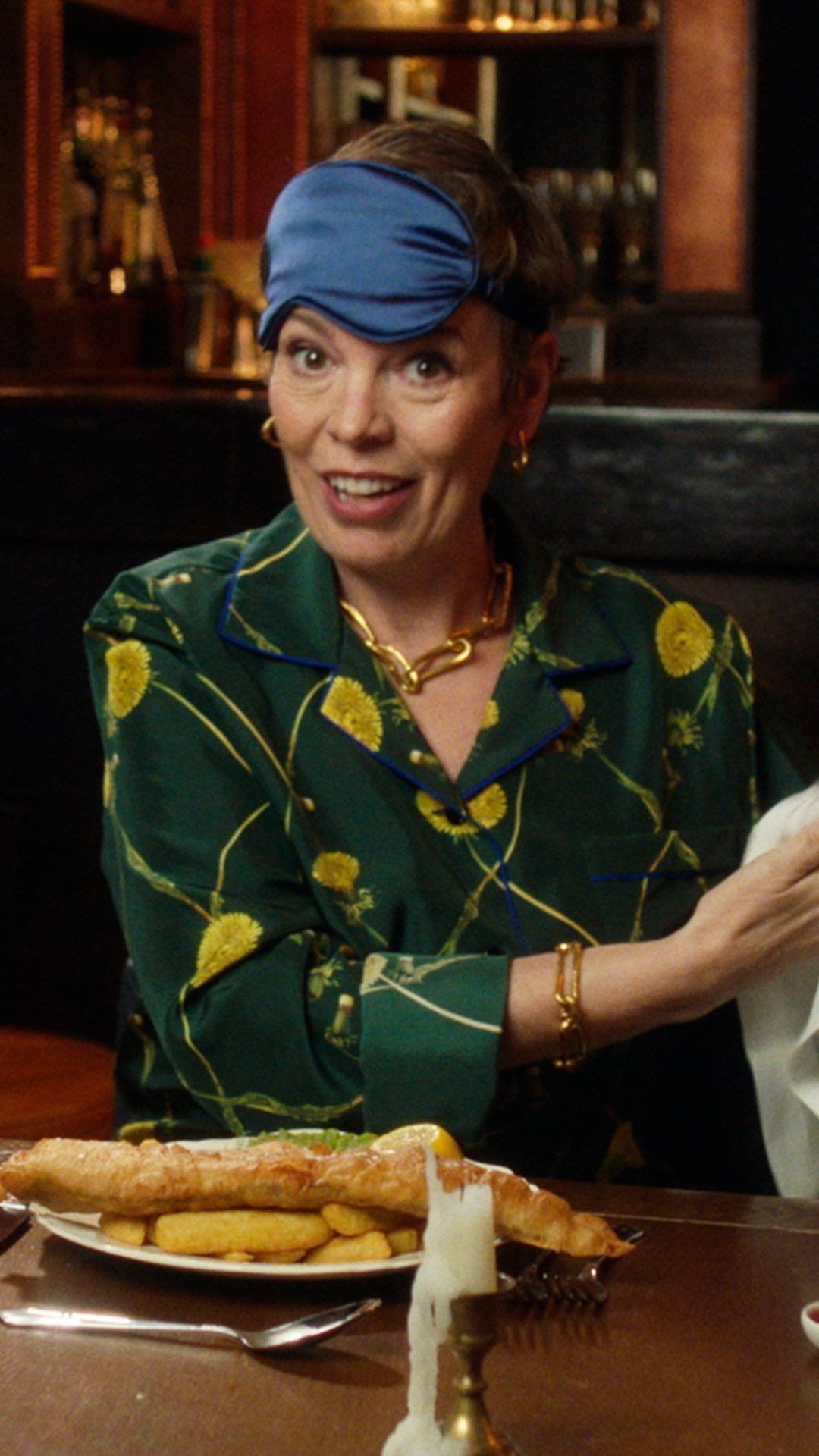 Woman in green floral shirt and blue headband sits at a table with a plate of fish and chips in a warmly lit restaurant setting.