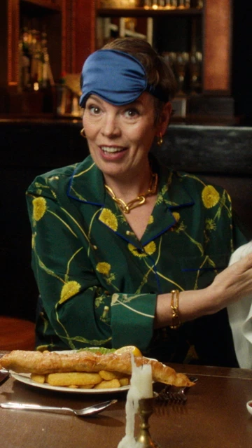 Woman in green floral shirt and blue headband sits at a table with a plate of fish and chips in a warmly lit restaurant setting.