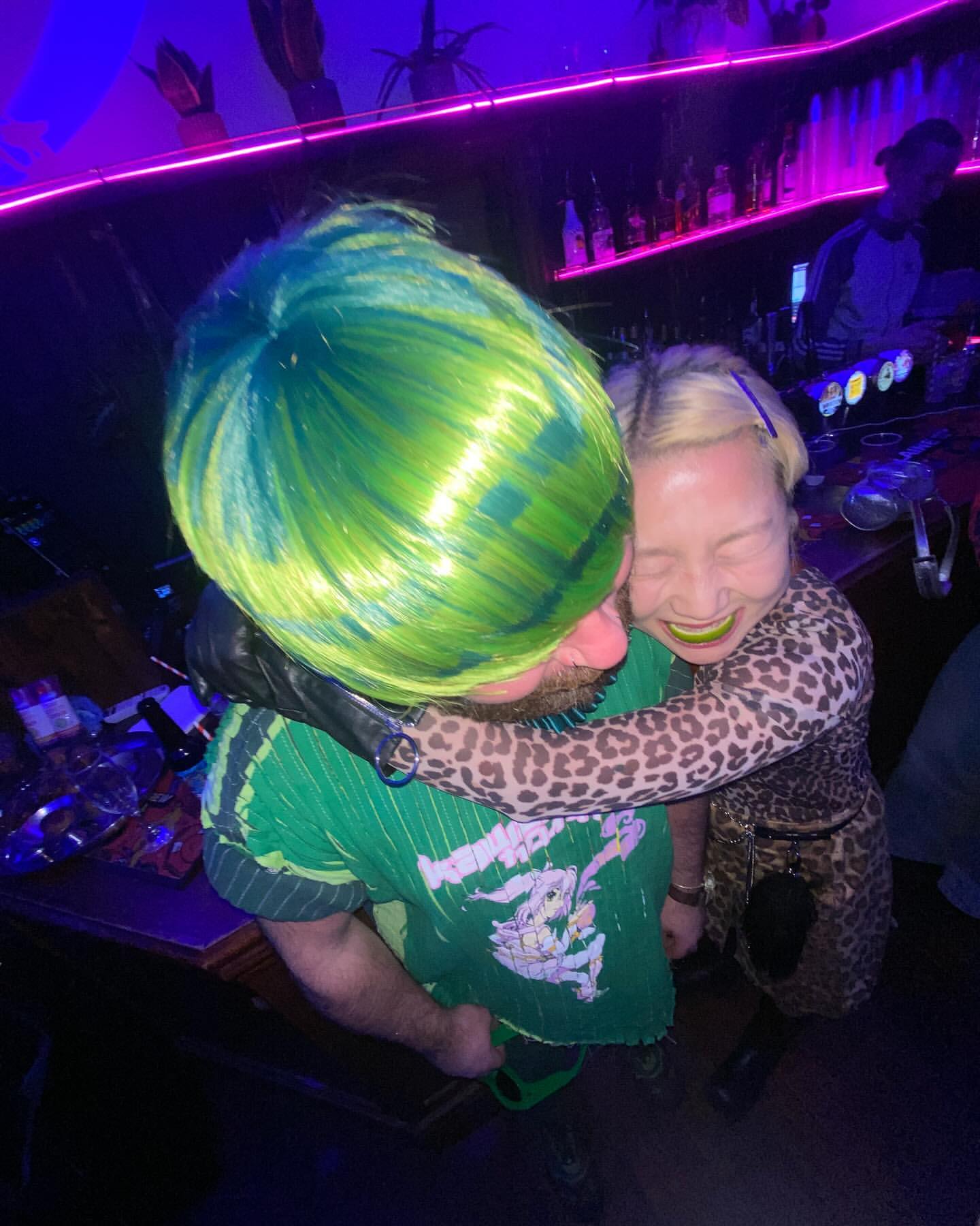 Two adults, one with green hair, embrace at a bar with purple lighting, leopard print dress, and green shirt in a candid moment.