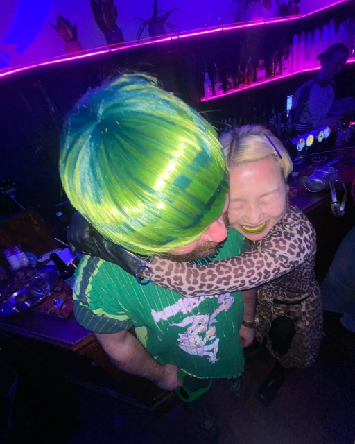 Two adults, one with green hair, embrace at a bar with purple lighting, leopard print dress, and green shirt in a candid moment.
