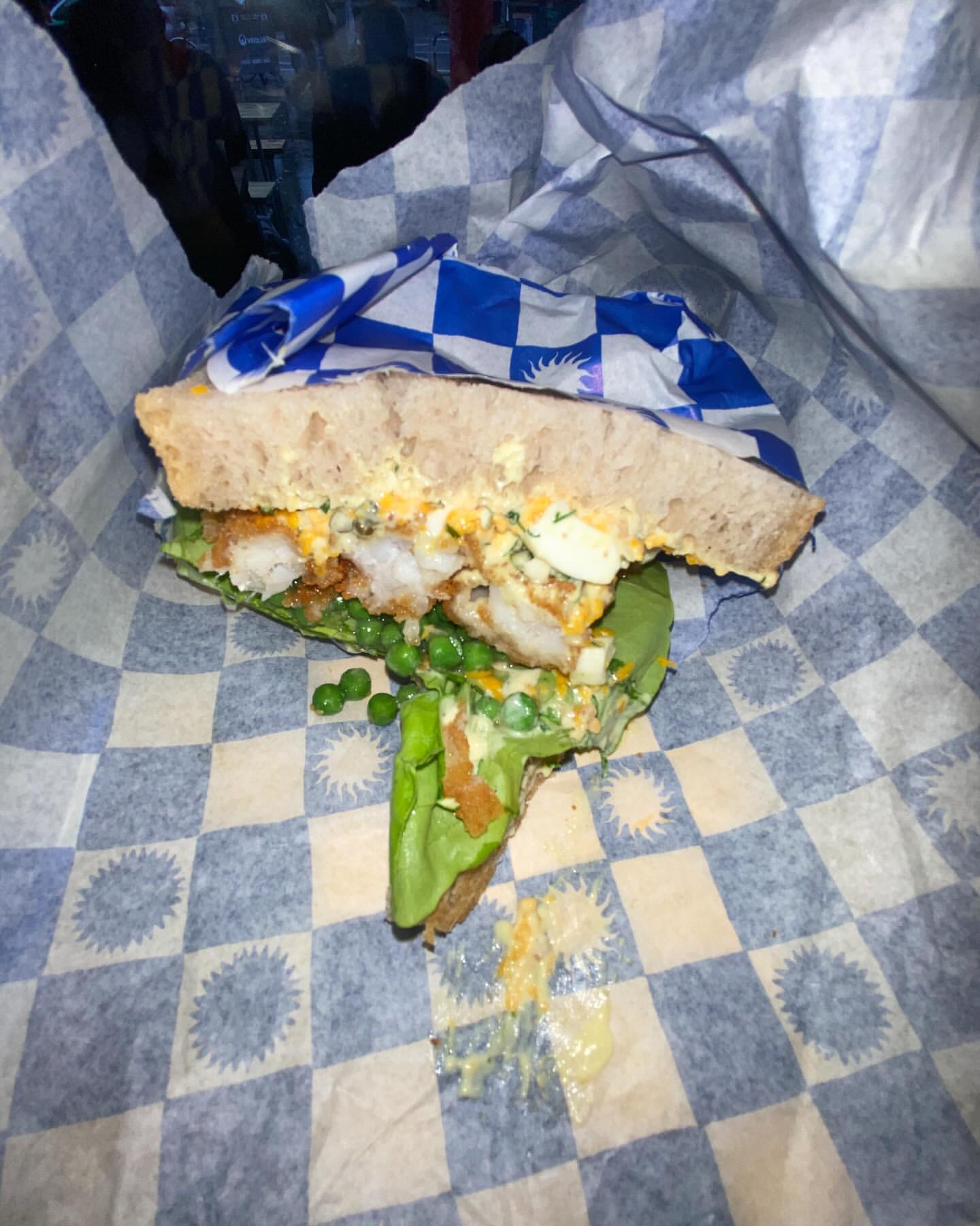 Sandwich featuring bread, lettuce, peas, and shrimp served on a blue and white checkered paper liner.