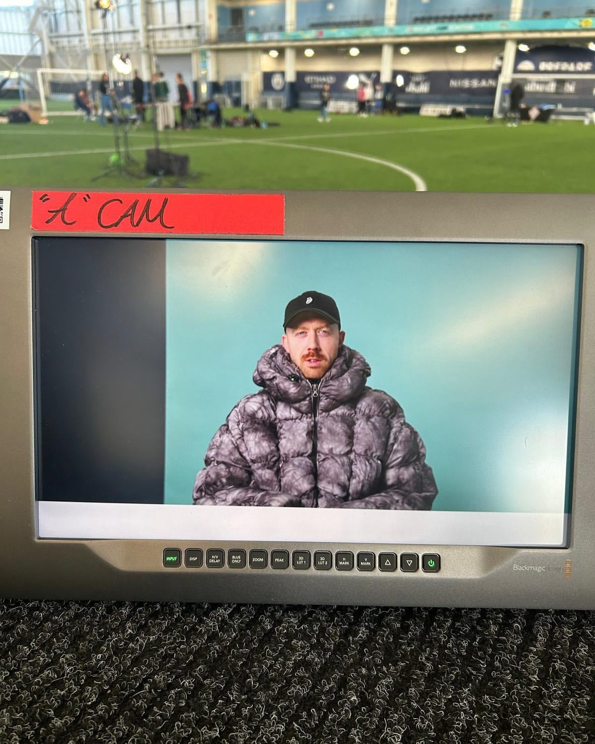 Man in a puffer jacket seen on a monitor screen in an indoor sports complex, with a camera crew and a green sports field behind.