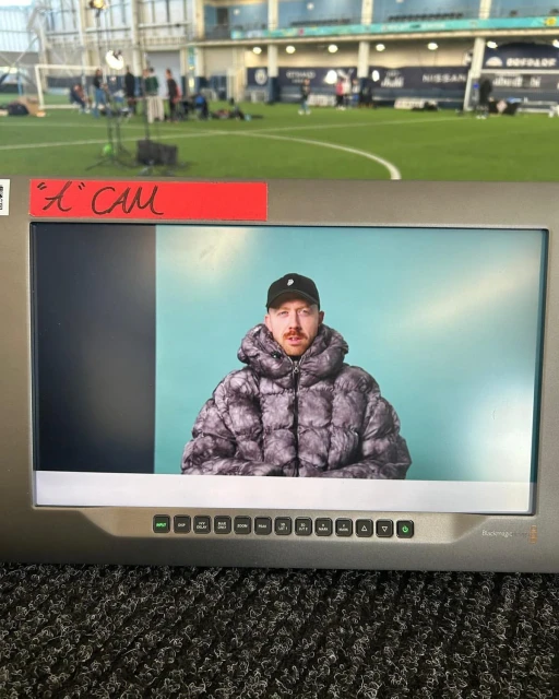 Man in a puffer jacket seen on a monitor screen in an indoor sports complex, with a camera crew and a green sports field behind.