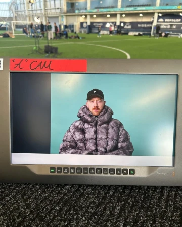Man in a puffer jacket seen on a monitor screen in an indoor sports complex, with a camera crew and a green sports field behind.