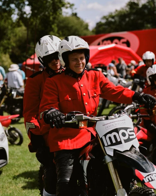 Two individuals in red uniform ride a motorcycle on a grassy field, surrounded by onlookers and tents in the background, bright daylight.