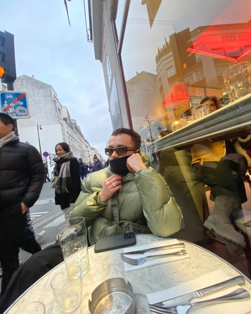 Adult male in an olive-green puffer jacket seated at a cafe table outdoors in an urban setting, seen through the glass, with city street background.