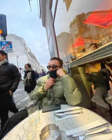 Adult male in an olive-green puffer jacket seated at a cafe table outdoors in an urban setting, seen through the glass, with city street background.