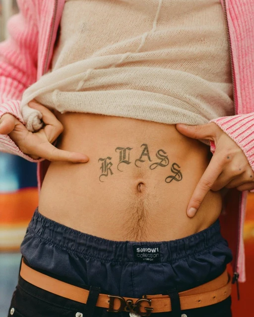 Close-up shot of a person's torso with a 'KLASS' tattoo above their navel, fingers pointing at the tattoo, wearing casual clothing.