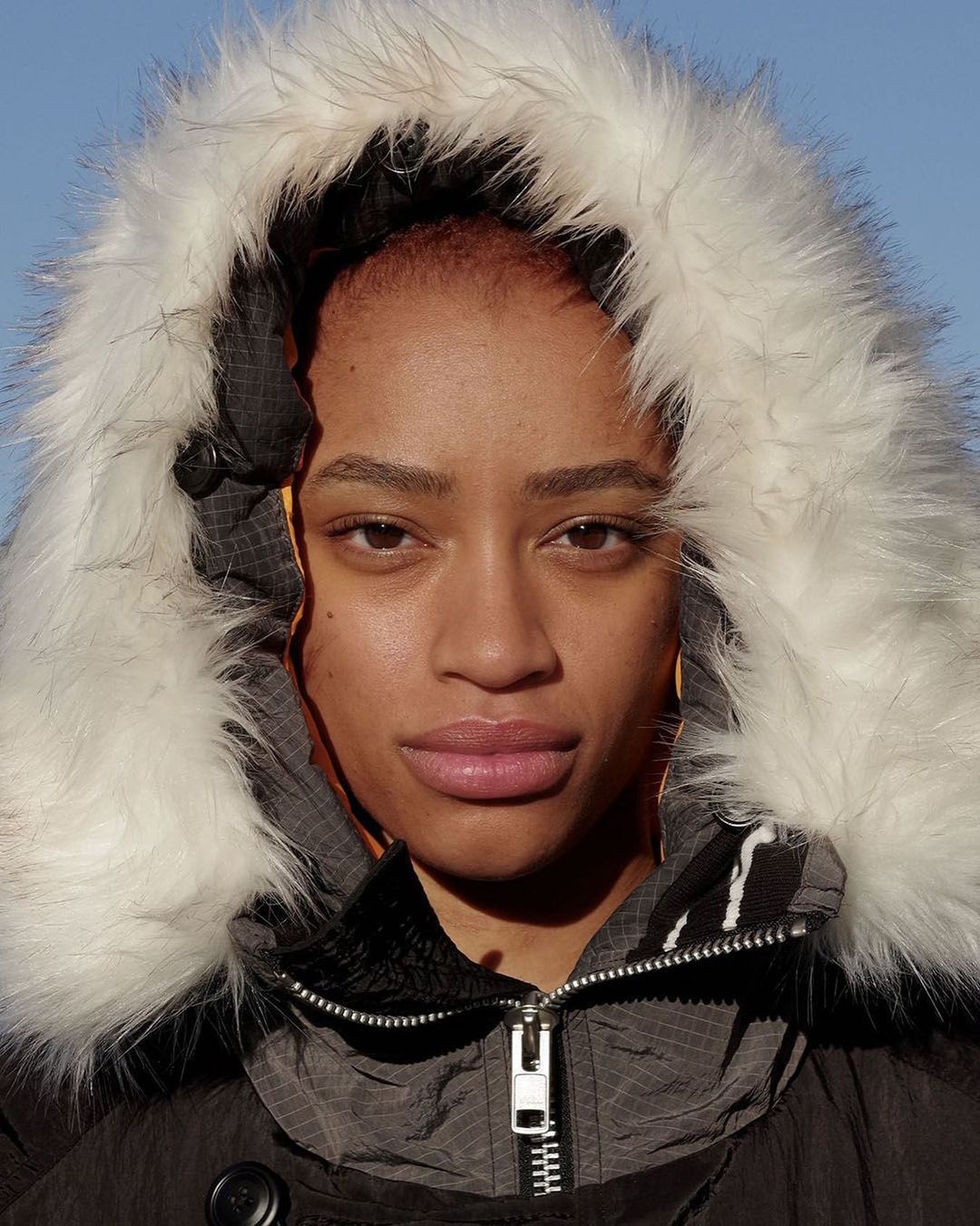 Close-up portrait of an adult female wearing a puffy winter coat with a white fur-trimmed hood against a clear blue sky.