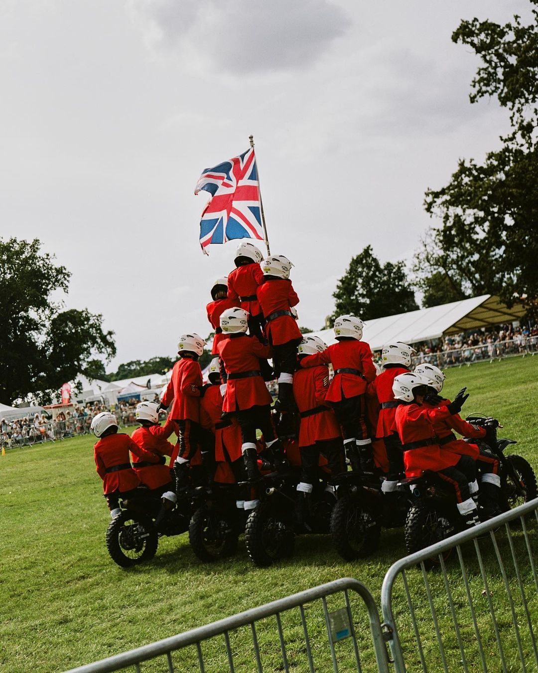 Acrobatic display featuring a large group of people on motorcycles forming a pyramid on green grass beneath the Union Jack.