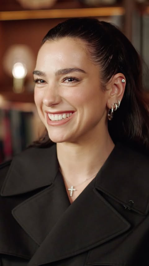Close-up of a smiling adult woman with dark hair wearing a black coat and cross necklace in an indoor setting with warm lighting.