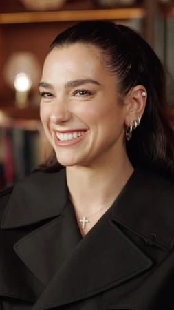 Close-up of a smiling adult woman with dark hair wearing a black coat and cross necklace in an indoor setting with warm lighting.