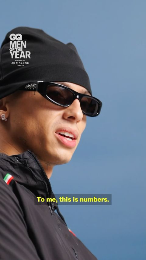 Portrait of a man wearing a black beanie and sunglasses, with a jacket and blue background, visible text indicates a GQ Men of the Year award.
