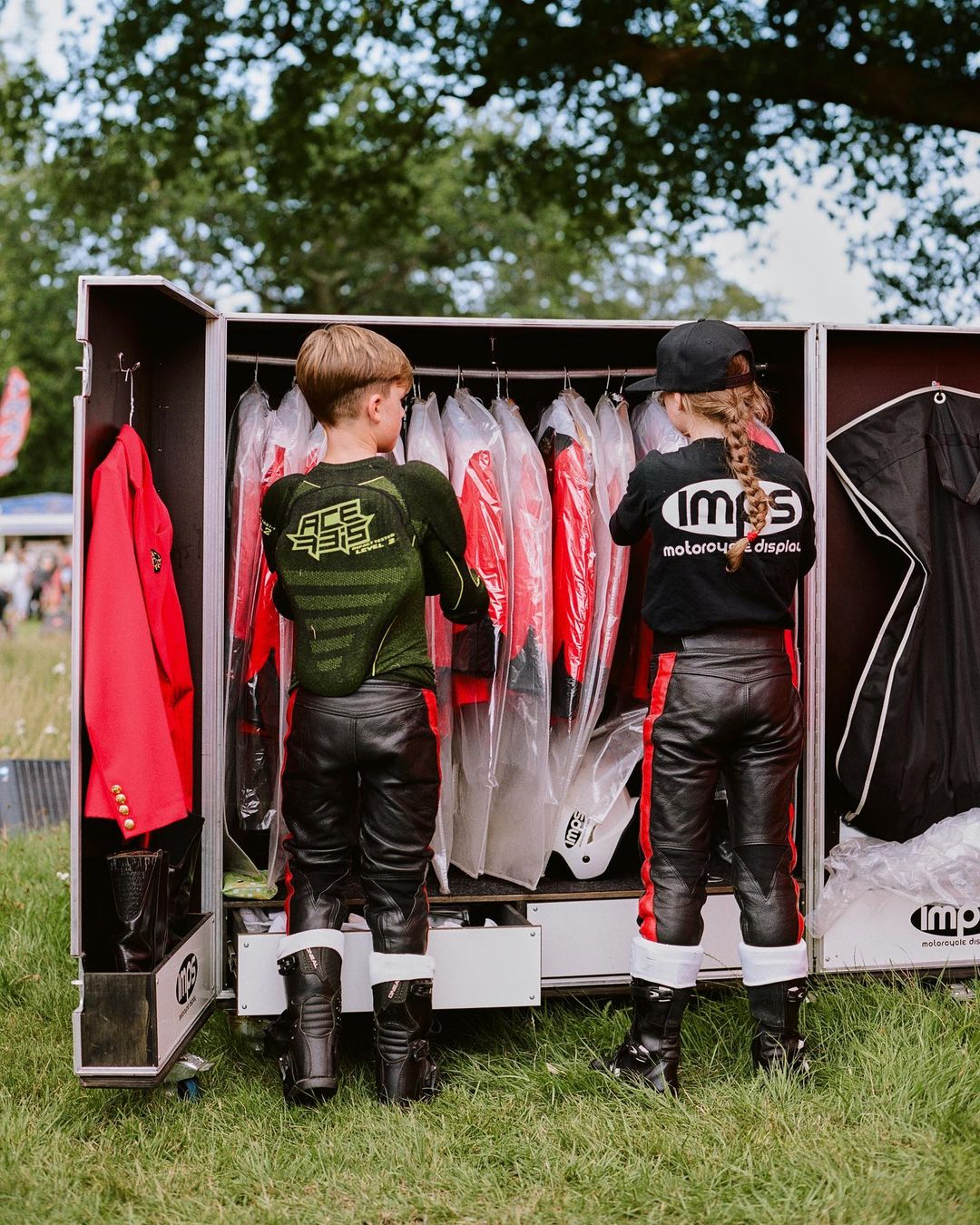 Two children in racing attire stand by a portable wardrobe with hanging racing suits, set on a grassy lawn with trees in the background.
