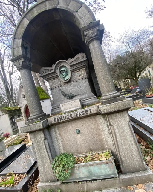 Ornate grey stone monument in a cemetery with a bronze portrait, decorative carvings, and surrounding greenery on an overcast day.