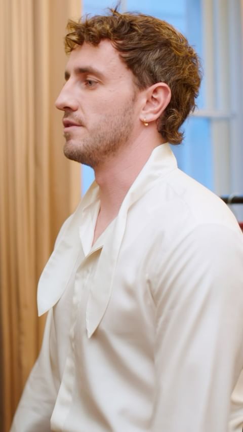 Portrait of an adult male in a white shirt with a collar, standing indoors near a window with a partially visible curtain.