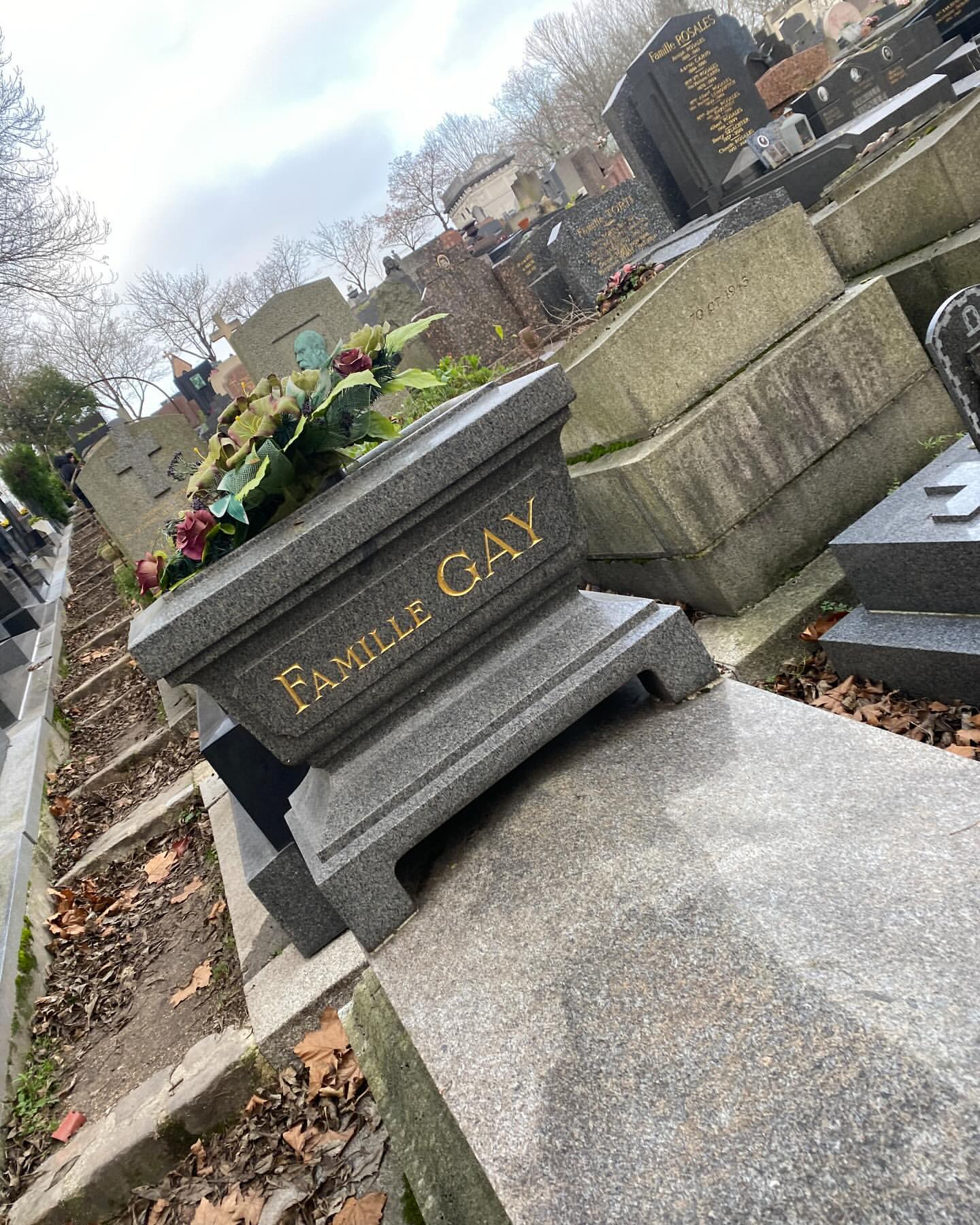 Tilted close-up on a 'Famille Gay' gravestone in a cemetery on a cloudy day. A flower arrangement sits on the tombstone.