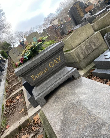 Tilted close-up on a 'Famille Gay' gravestone in a cemetery on a cloudy day. A flower arrangement sits on the tombstone.