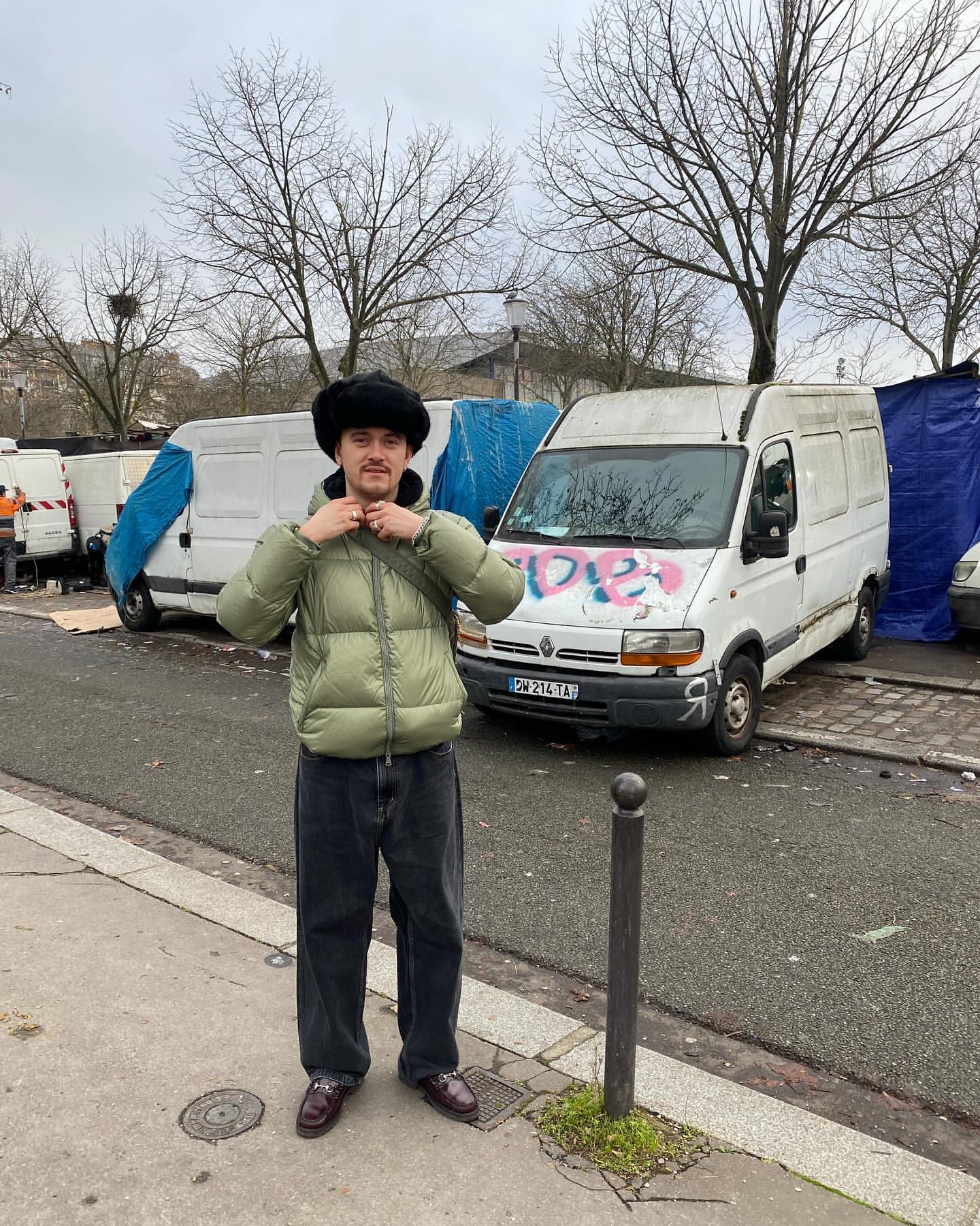A person standing on a city street wearing a green puffer jacket and black fur hat with white vans and bare trees in the background.