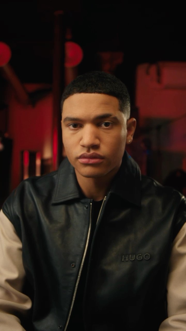 Close-up of a young adult male with short hair wearing a black and beige Hugo varsity jacket indoors with red lighting in the background.