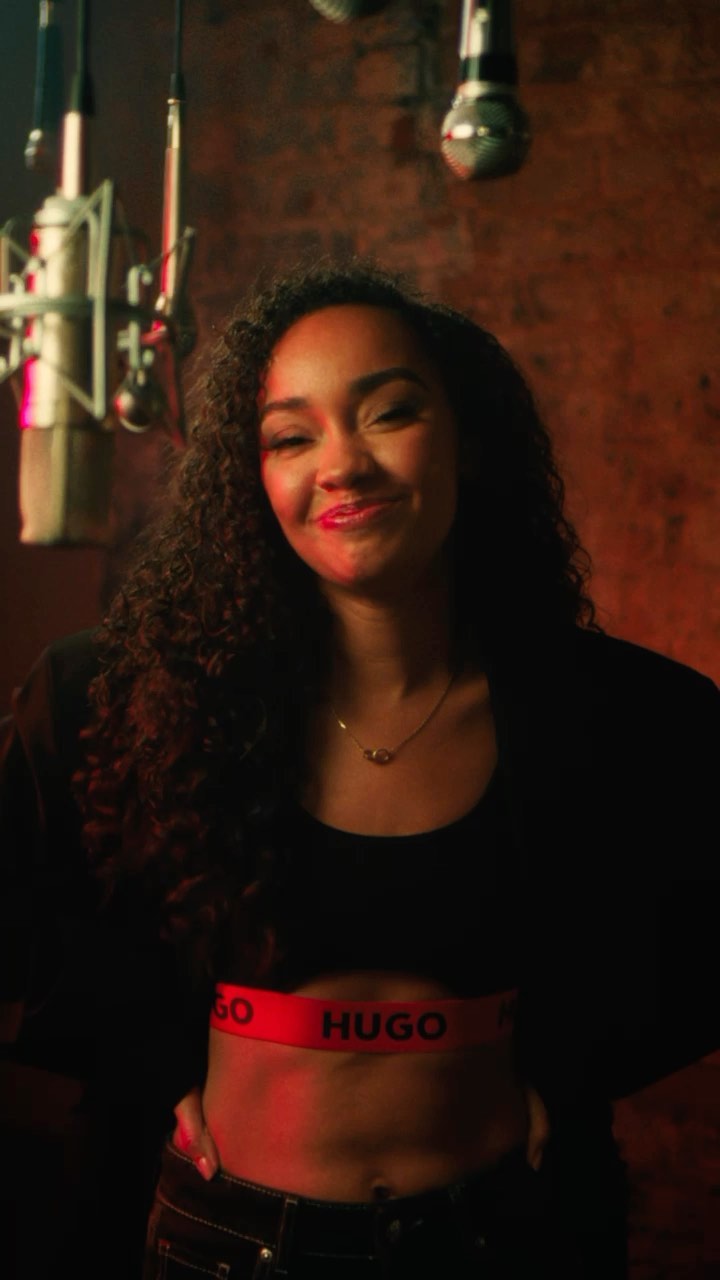 A stylish woman in a black top poses in a warm-toned room with visible studio microphones and brick background, creating an intimate, musical atmosphere.