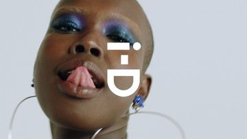 Close-up of an adult woman with bronze skin and blue eyeshadow, sticking out her tongue, posing in front of a light gray background.