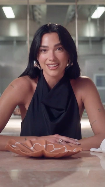 Brunette woman with hoop earrings sits at a table with a ceramic plate, wearing a black top in an indoor kitchen environment.