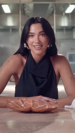 Brunette woman with hoop earrings sits at a table with a ceramic plate, wearing a black top in an indoor kitchen environment.