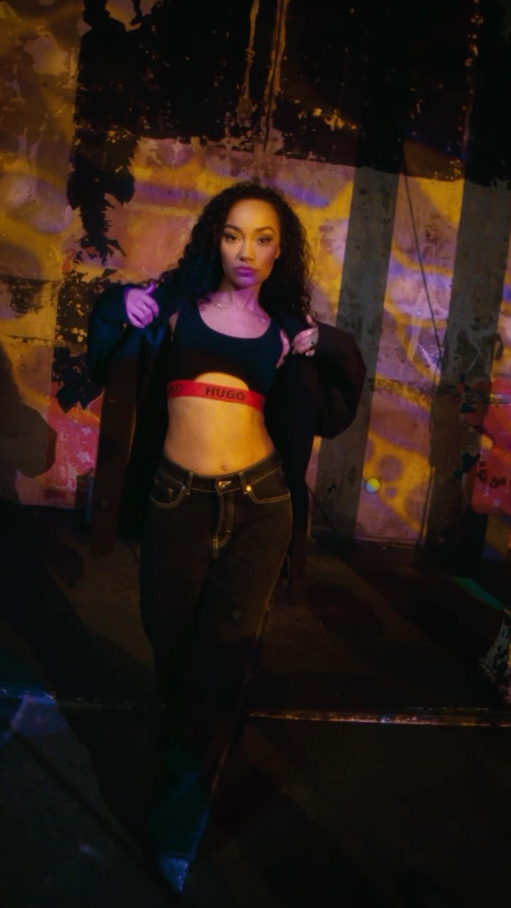A woman with long curly hair poses indoors wearing a black jacket, bra top, and jeans, against a graffiti-style background in dramatic lighting.