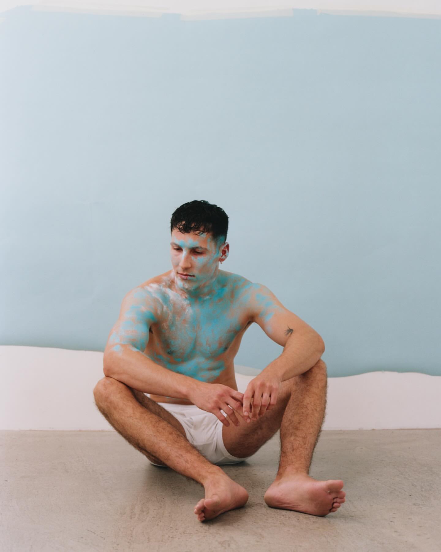 Shirtless man sitting with blue paint on his skin in front of a light blue and white wall, looking off to the side. 