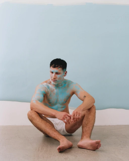 Shirtless man sitting with blue paint on his skin in front of a light blue and white wall, looking off to the side. 