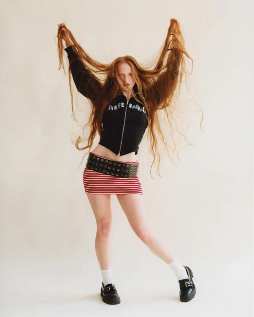 Full-length studio shot of a young adult woman with long red hair, wearing a black jacket and a striped mini skirt. She poses playfully.