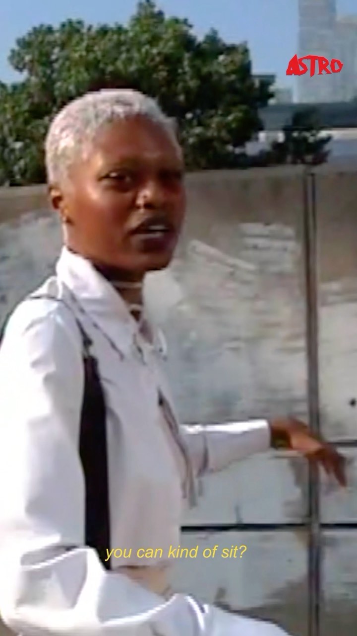Adult woman with short platinum hair sits outside near a concrete wall with a cityscape and blue sky in the distance, wearing a white jacket.