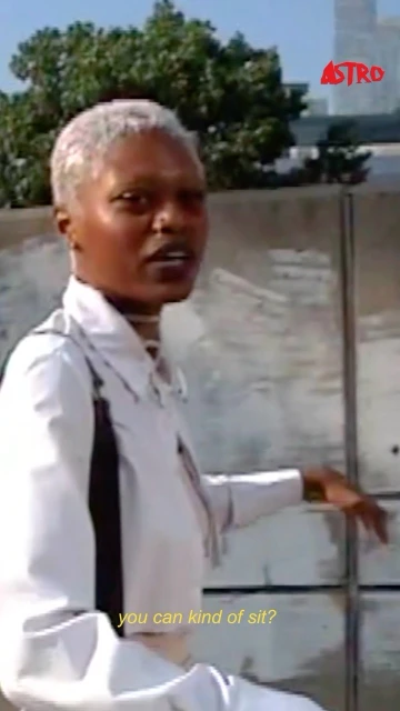 Adult woman with short platinum hair sits outside near a concrete wall with a cityscape and blue sky in the distance, wearing a white jacket.