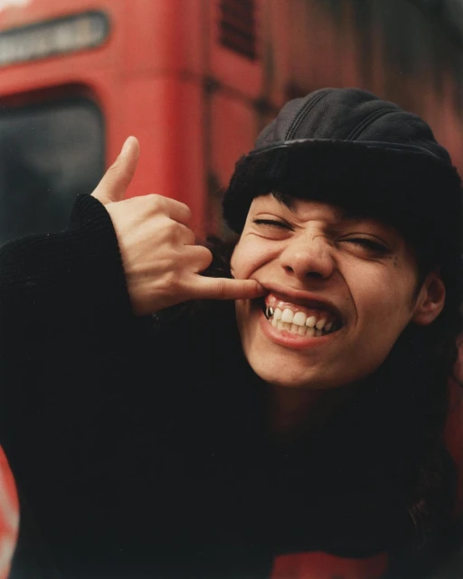 Close-up of a woman with dark hair, wearing a black hat and sweater, making a 'call me' gesture with her hand. Red metal background.