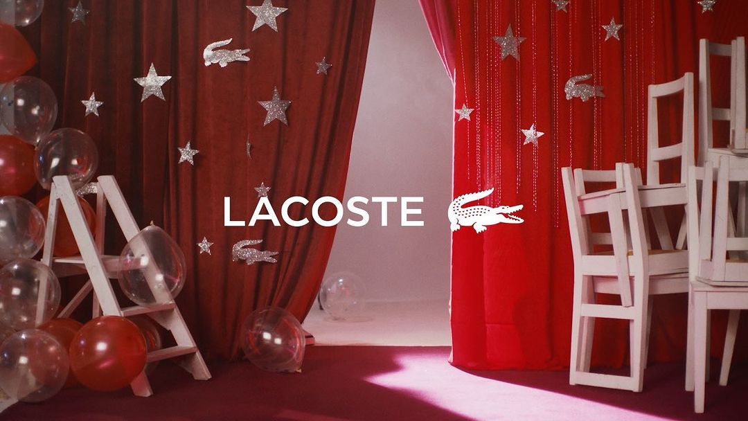 Lacoste ad featuring red balloons, a white ladder, stacked chairs, red curtains, and star decorations surrounding the Lacoste logo.