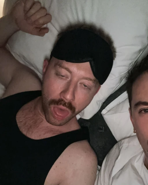 Two adult males are lying side-by-side on a white bed. One man is wearing a sleep mask and looking down; the other is partially visible.