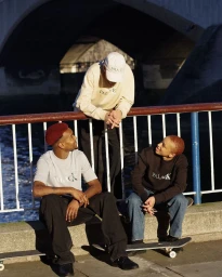 Three young skateboarders wearing branded sweatshirts gathered outdoors near a bridge by the water, photographed by Tom Ivin.