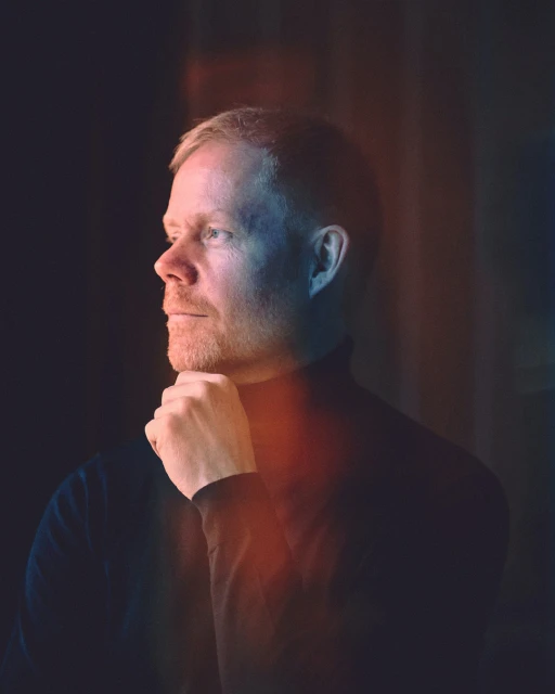 Portrait of a blond man with a beard and blue eyes, wearing a dark turtleneck, looking to the left, hand on chin, in a dimly lit setting.