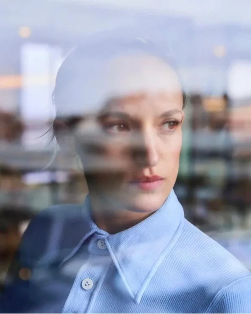 Portrait of a woman with short hair, wearing a blue shirt and looking to the side, with a window reflection overlaying the scene.