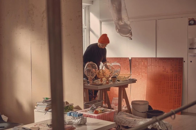 Adult working at a table with glass vases and bowls in an artist's studio with canvases and buckets, in a room with natural light.