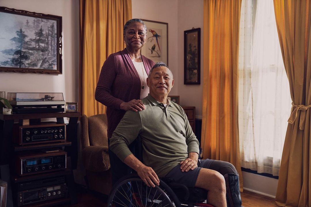 Elderly Asian man in wheelchair with arm around African-American woman in living room, featuring vintage audio equipment and warm lighting.