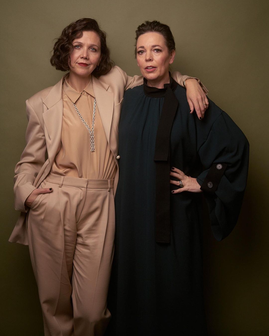Two adult females, Maggie Gyllenhaal and Olivia Colman, pose in a studio setting. Gyllenhaal wears a suit, and Colman wears a dark green dress.