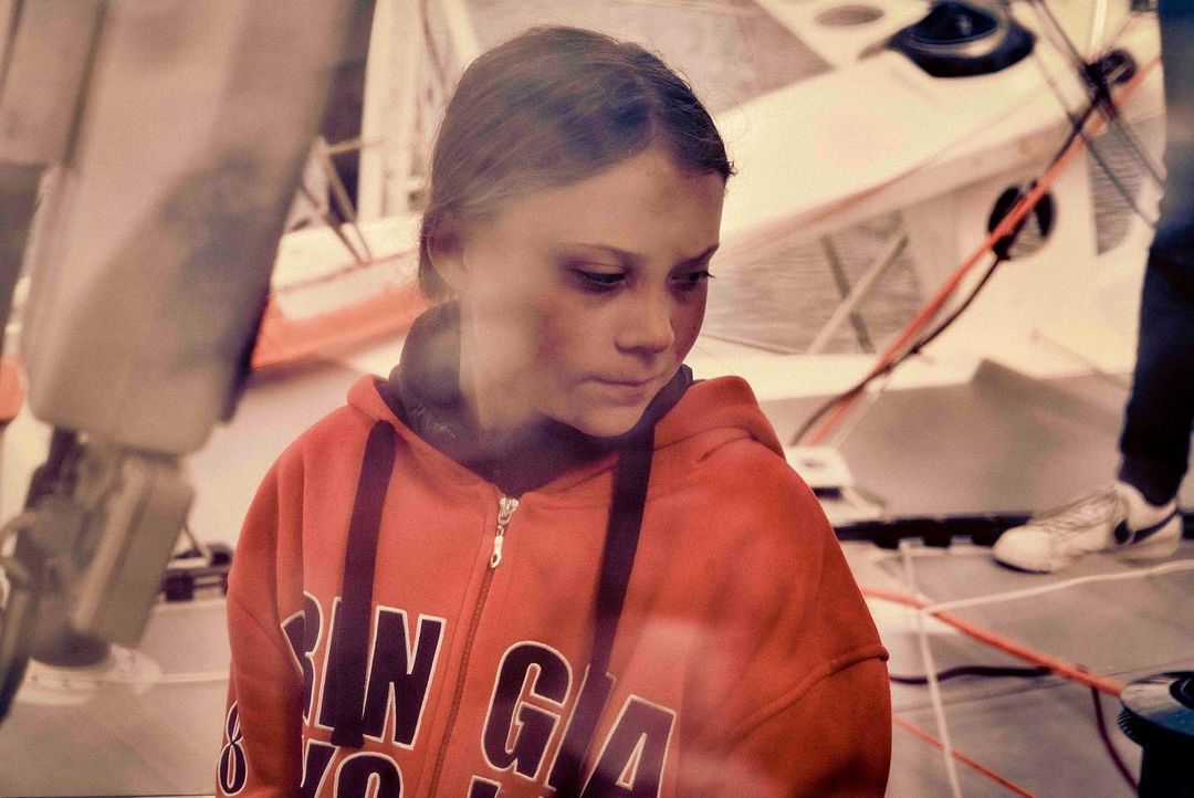 Teenage girl in a red hooded sweatshirt looks down in an indoor location with miscellaneous equipment visible in the background of the frame.