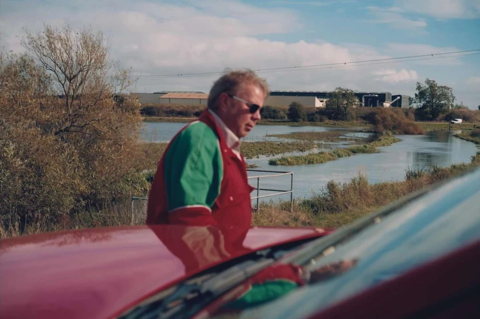 Adult male in sunglasses and red jacket stands near a red car in a rural landscape with lake and buildings on a sunny day.