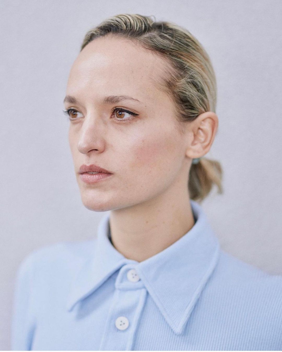 Close-up portrait of a fair-skinned woman with light blonde hair, wearing a blue button-up shirt, against a light grey background.