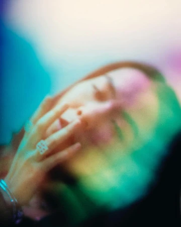 Close-up portrait of a woman with her hand near her face, bathed in colorful prismatic light flares against a dark background.