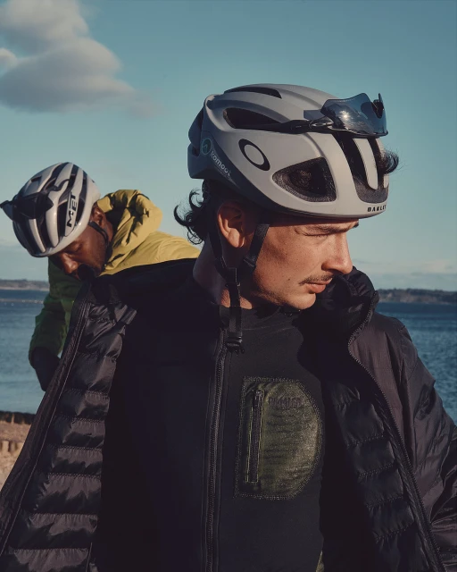 Two cyclists wearing helmets and jackets are shown outdoors against a blue sky with a body of water in the distance. The focus is on the cyclist in front.