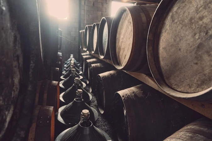 Stacks of wooden barrels and glass demijohns line the walls in a dimly lit wine cellar, showing vintage winemaking process.