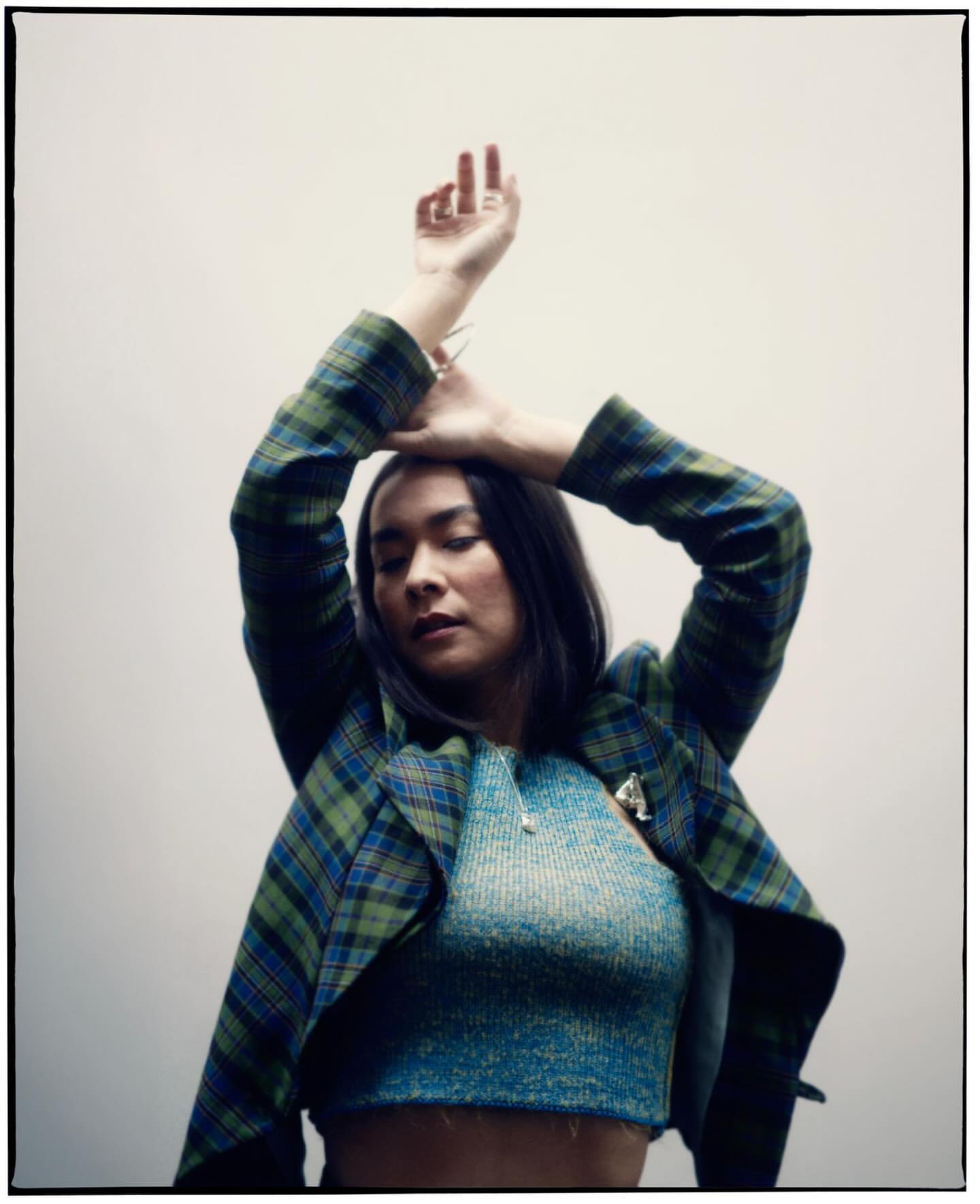 Portrait of a young woman with her arms raised, wearing a blue and green plaid jacket and blue shirt, against a light grey background.